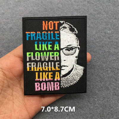 Embroidered patch with text 'Not fragile like a flower, fragile like a bomb' held by a hand on a gray background.