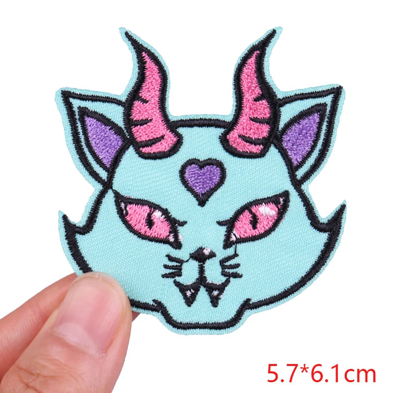 Embroidered patch of a cat-like creature with pink horns and eyes, held by a hand on a white background.