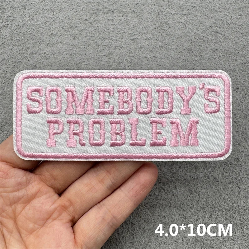 Hand holding a pink and white embroidered Somebody’s Problem iron-on patch approximately 4x10cm on a gray background