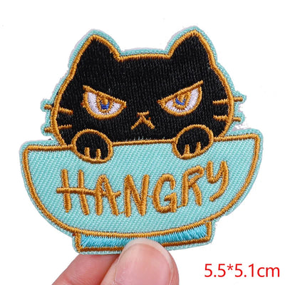 Embroidered patch of a black cat in a bowl with 'HANGRY' text, held by a hand.