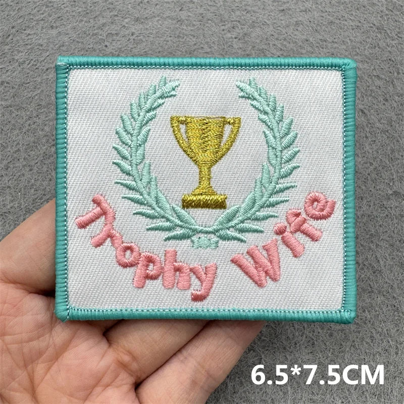 Embroidered patch with 'Trophy Wife' text held by a hand on a gray background