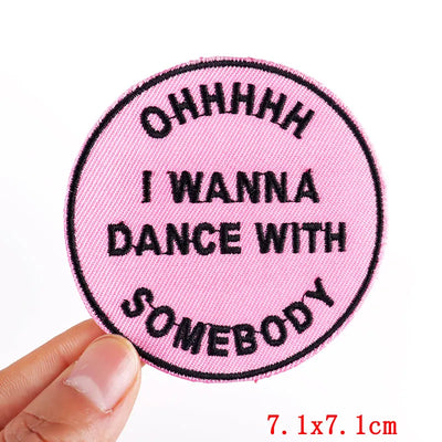 Pink patch with black text 'OHHHHH I WANNA DANCE WITH SOMEBODY' held by a hand on a white background.