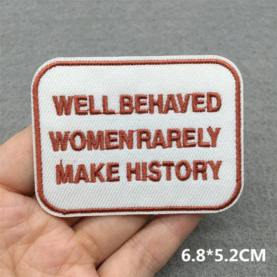 Patch with text 'WELL BEHAVED WOMEN RARELY MAKE HISTORY' held in a hand against a gray background.