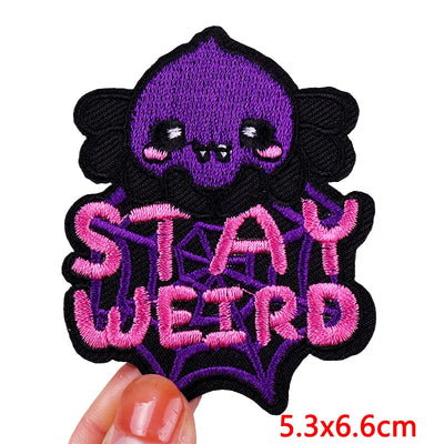 Embroidered patch with a purple skull and 'Stay Weird' text held by a hand.