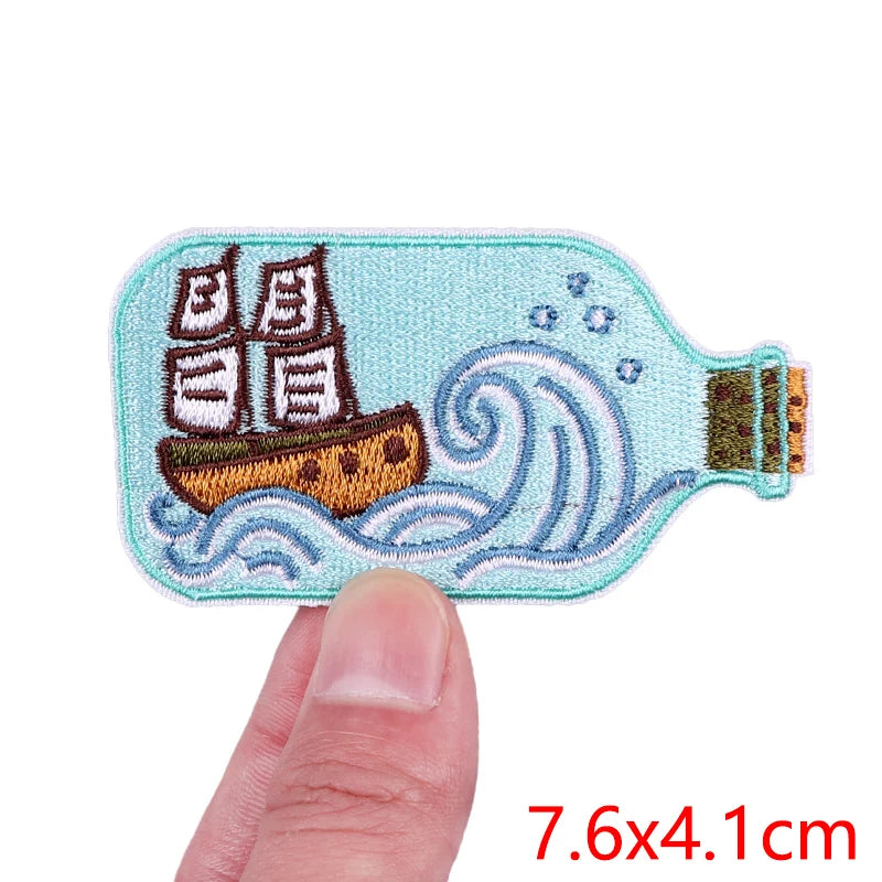 Embroidered patch of a ship in a bottle held between fingers with size label '7.6x4.1cm'.