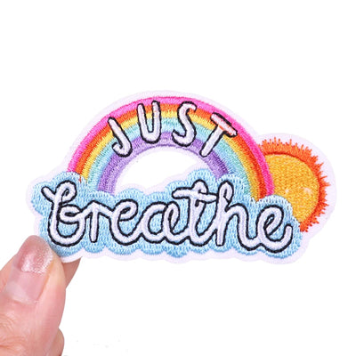 Colorful patch with rainbow and 'Just Breathe' text held by a hand on a white background