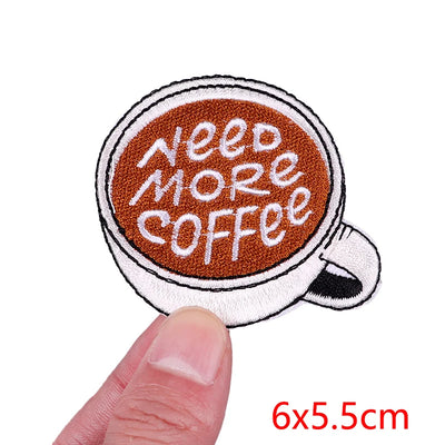 Patch of a coffee cup with 'Need More Coffee' text held by a finger, on a white background.
