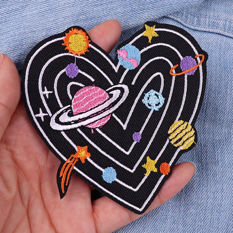 Heart-shaped patch with space-themed design held in a hand against a denim background