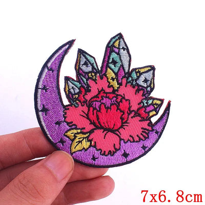 Embroidered patch with floral design and purple crescent moon held by a hand, on a white background.