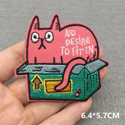 Embroidered patch of a pink cat in a box with text on a gray background