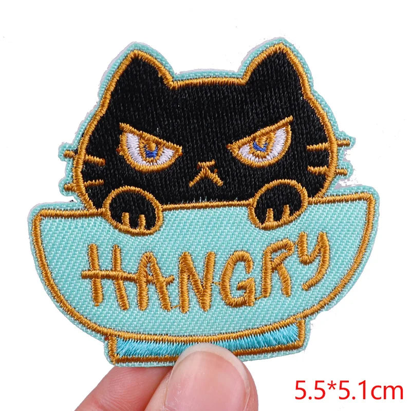Embroidered patch of a black cat in a bowl with 'HANGRY' text, held by a hand.