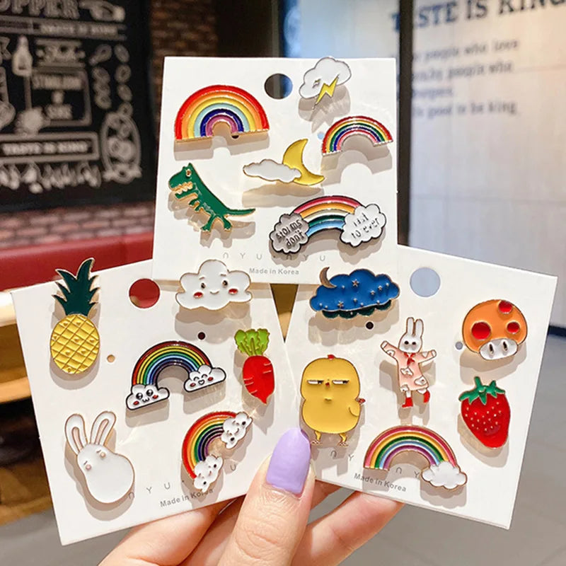 Three cards with colorful enamel pins featuring rainbows, animals, and other designs held by a hand.