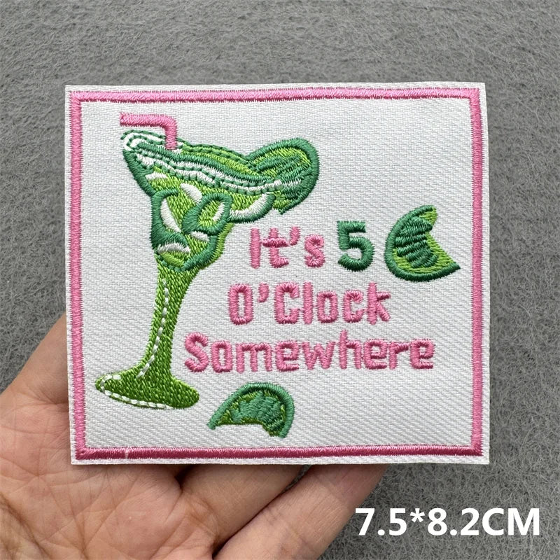 Embroidered patch with a cocktail design and text 'It's 5 O'Clock Somewhere' held by a hand on a gray background.