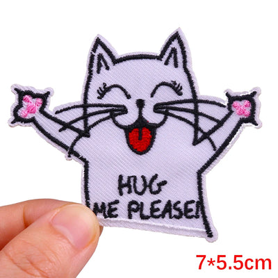 Embroidered patch of a cat with 'Hug Me Please!' text held by a hand on a white background.