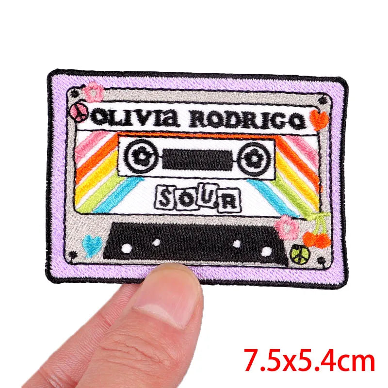 Embroidered patch of a cassette tape with 'Olivia Rodrigo Sour' text, held by a finger for scale.
