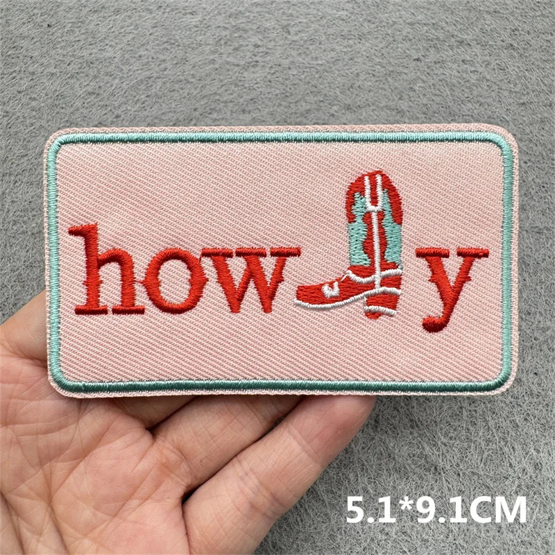 Embroidered patch with 'howdy' text and a boot design held by a hand on a gray background.