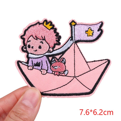Embroidered patch of a character with pink hair on a pink paper boat, held by a hand.