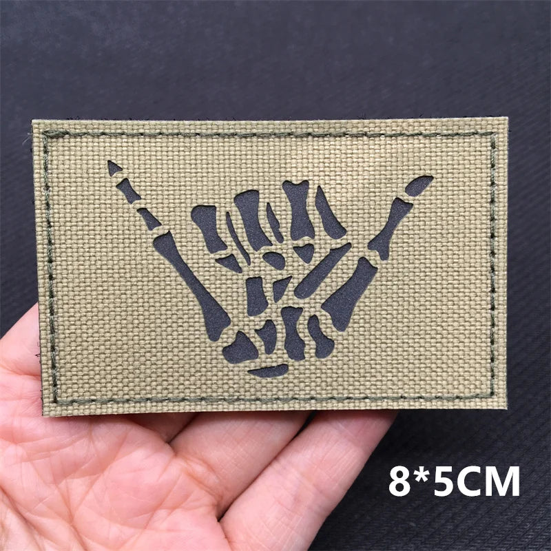 Kaki green patch with black skeleton hand design held in a hand against a dark background