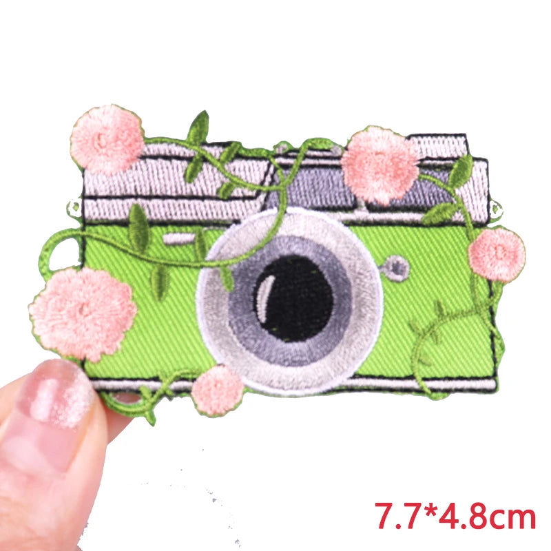 Hand holding a floral vintage camera embroidered iron-on patch featuring a green camera surrounded by pink flowers and trailing vines approximately 7.7x4.8cm on a white background