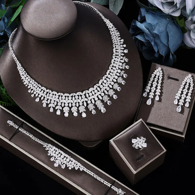 Silver jewelry set including necklace, bracelet, earrings, and ring on a dark surface with flowers in the background.