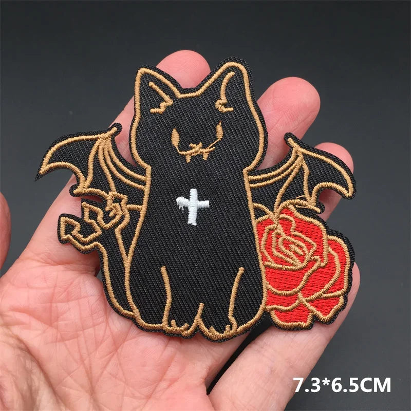 Black cat patch with bat wings and red rose held in a hand against a dark background