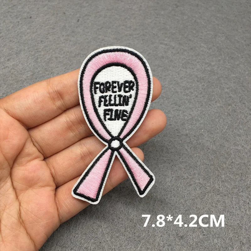 Pink ribbon patch with 'Forever Feelin' Fine' text held in a hand on a gray background