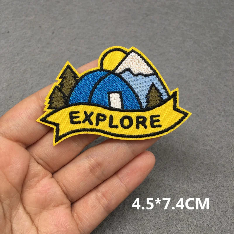 Embroidered patch with 'EXPLORE' text and camping design held in a hand on a gray background.