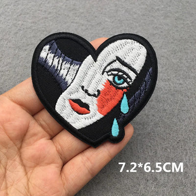 Heart-shaped patch with a stylized face design held in a hand on a gray background