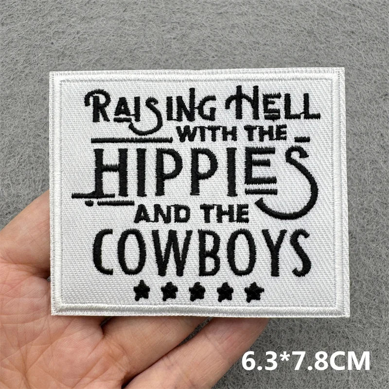 Hand holding a black and white embroidered Raising Hell with the Hippies and the Cowboys iron-on patch approximately 6.3x7.8cm on a gray background