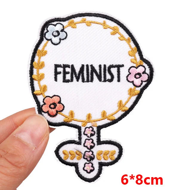 Hand holding a white floral feminist symbol embroidered iron-on patch with pink flowers and gold vine details approximately 6x8cm on a white background