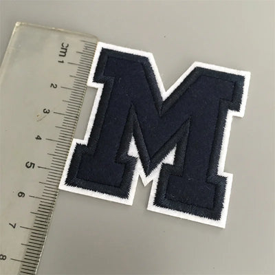 Navy blue letter 'M' patch next to a ruler on a gray background