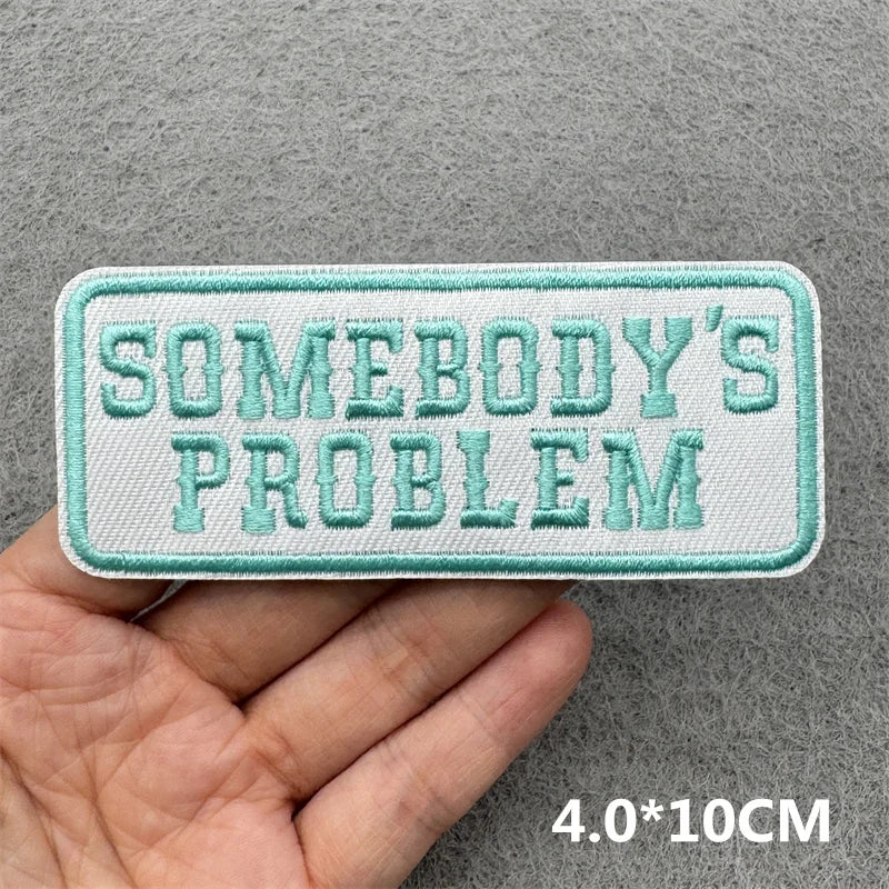 Hand holding a mint green and white embroidered Somebody’s Problem iron-on patch approximately 4x10cm on a gray background