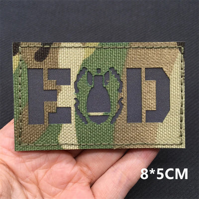 Explosive Ordnance Disposal Camouflage patch with 'EOD' and bomb symbol held in a hand, on a dark background.