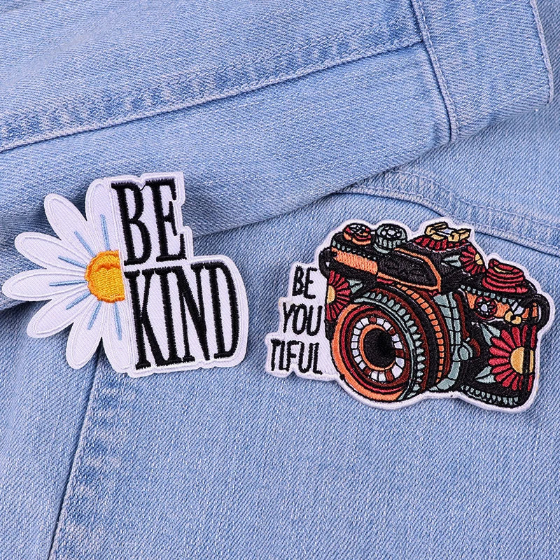 Two patches on a denim jacket: one with 'BE KIND' text and a daisy, the other with a camera and 'BE YOU TIFUL' text.