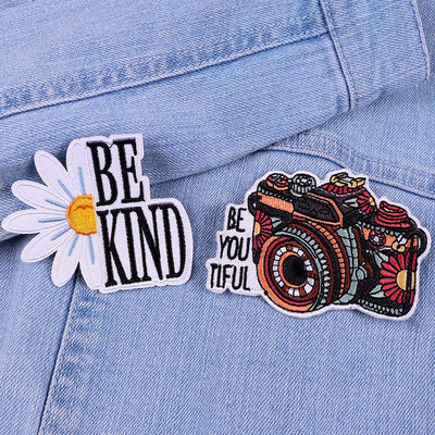 Two patches on a denim jacket: one with 'BE KIND' text and a daisy, the other with a camera and 'BE YOU TIFUL' text.