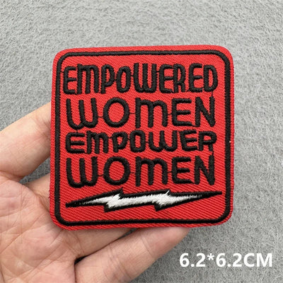 Red patch with 'EMPOWERED WOMEN EMPOWER WOMEN' text held by a hand on a gray background