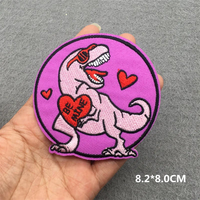 Embroidered patch with a dinosaur holding a heart and 'Be Mine' text on a pink circle, held in a hand against a gray background.