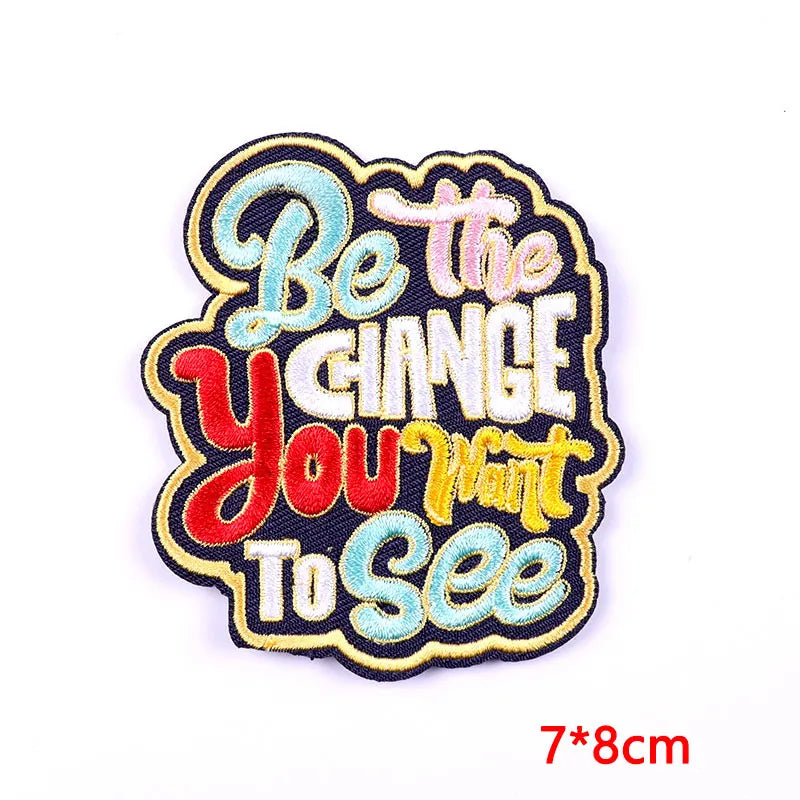 Be The Change You Want To See embroidered iron-on patch featuring sky blue, red, golden yellow, and white retro lettering on a black background, approximately 7x8cm, photographed on a white background.