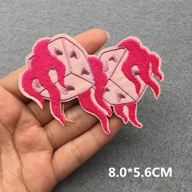 Two pink embroidered patches of a woman and a box held between fingers on a gray background.