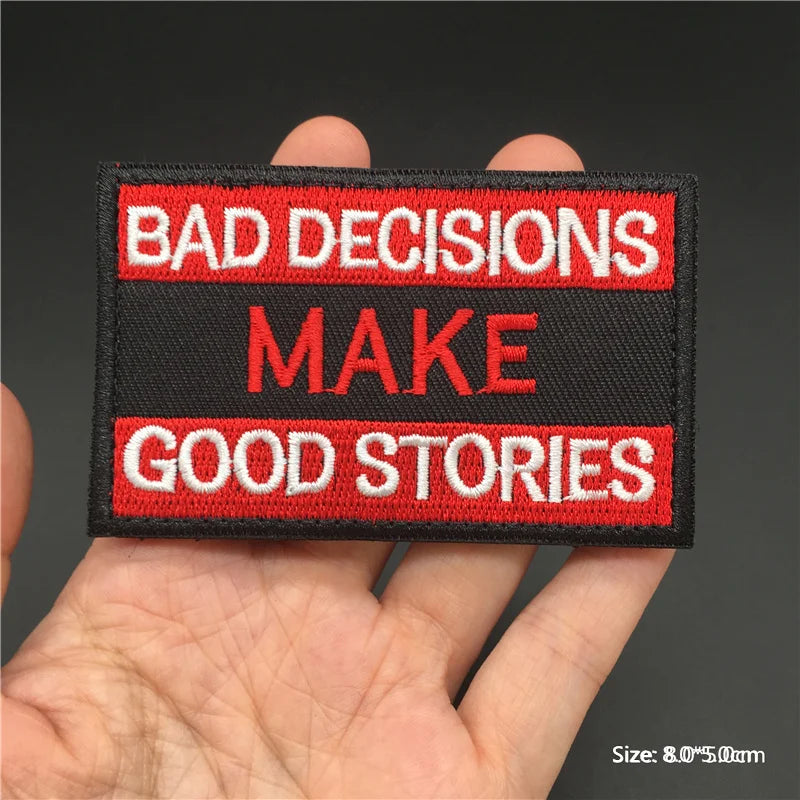 Hand holding a black red and white Bad Decisions Make Good Stories embroidered hook-and-loop patch approx 8x5cm on a dark background