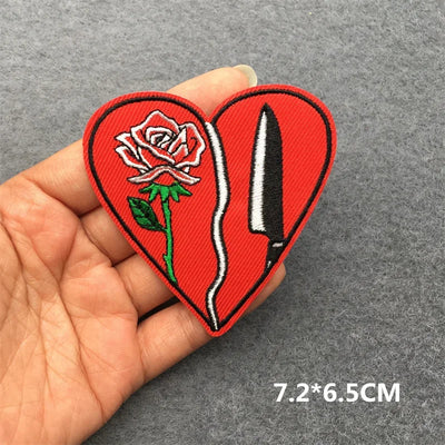 Heart-shaped patch with a rose and knife design held by a hand on a gray background