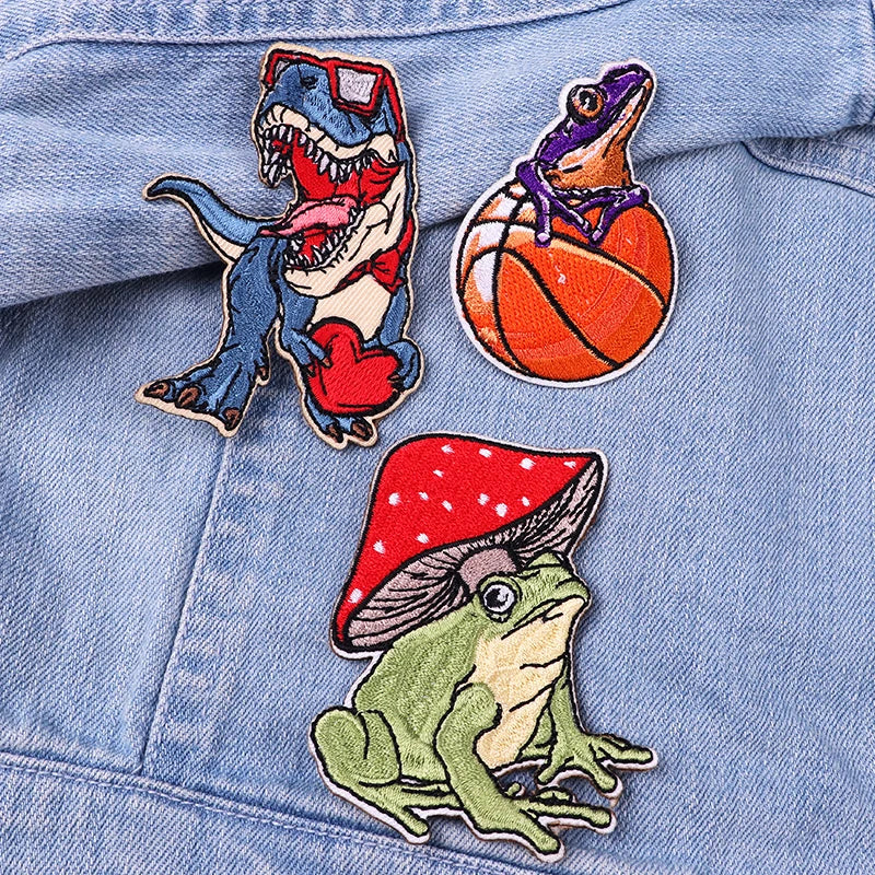 Three colorful patches on a denim jacket: a dinosaur, a frog with a basketball, and a mushroom with a frog.