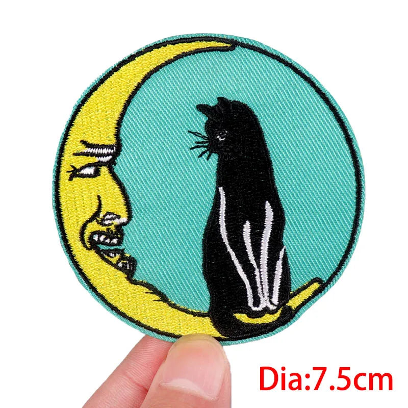 Embroidered patch with a black cat on a yellow crescent moon against a teal background, held by a hand.