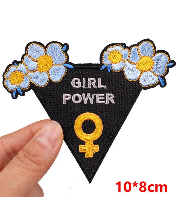 Hand holding a black triangular Girl Power embroidered iron-on patch with blue flowers and a yellow female symbol approximately 10x8cm on a white background