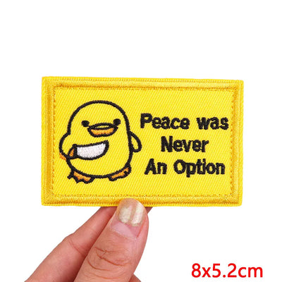 Yellow patch with cartoon duck and text 'Peace was Never An Option' held by a hand on a white background.