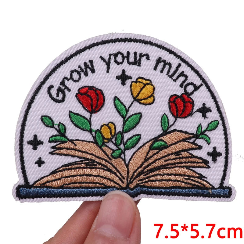 Embroidered patch with 'Grow your mind' text and flowers on an open book, held by a hand.