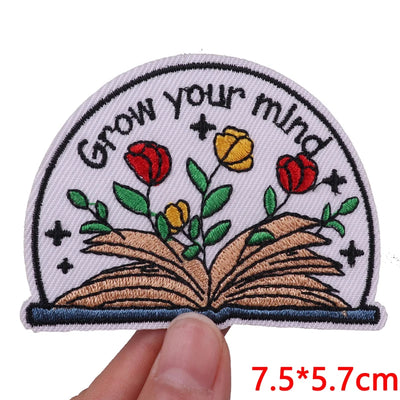 Embroidered patch with 'Grow your mind' text and flowers on an open book, held by a hand.