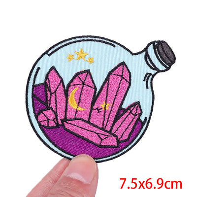 Embroidered patch of pink crystals in a bottle held by a hand, with dimensions 7.5x6.9cm.
