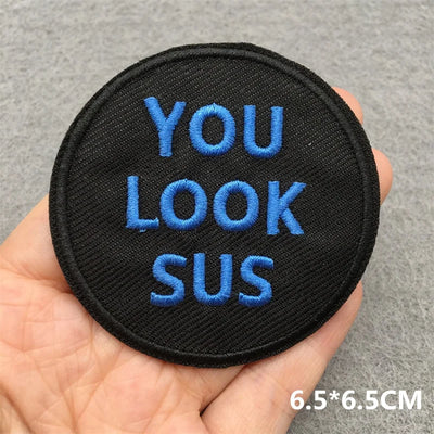 Black patch with blue text 'YOU LOOK SUS' held in a hand against a gray background