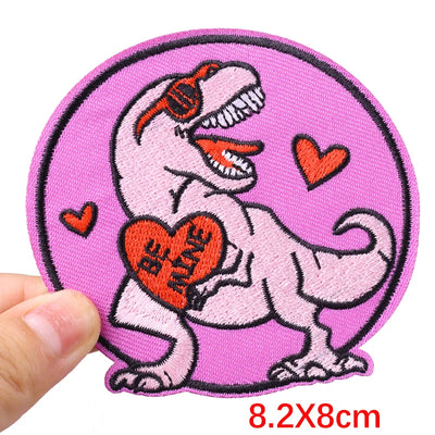 Embroidered patch of a dinosaur holding a heart with 'Be Alive' text on a pink background, held by a hand.